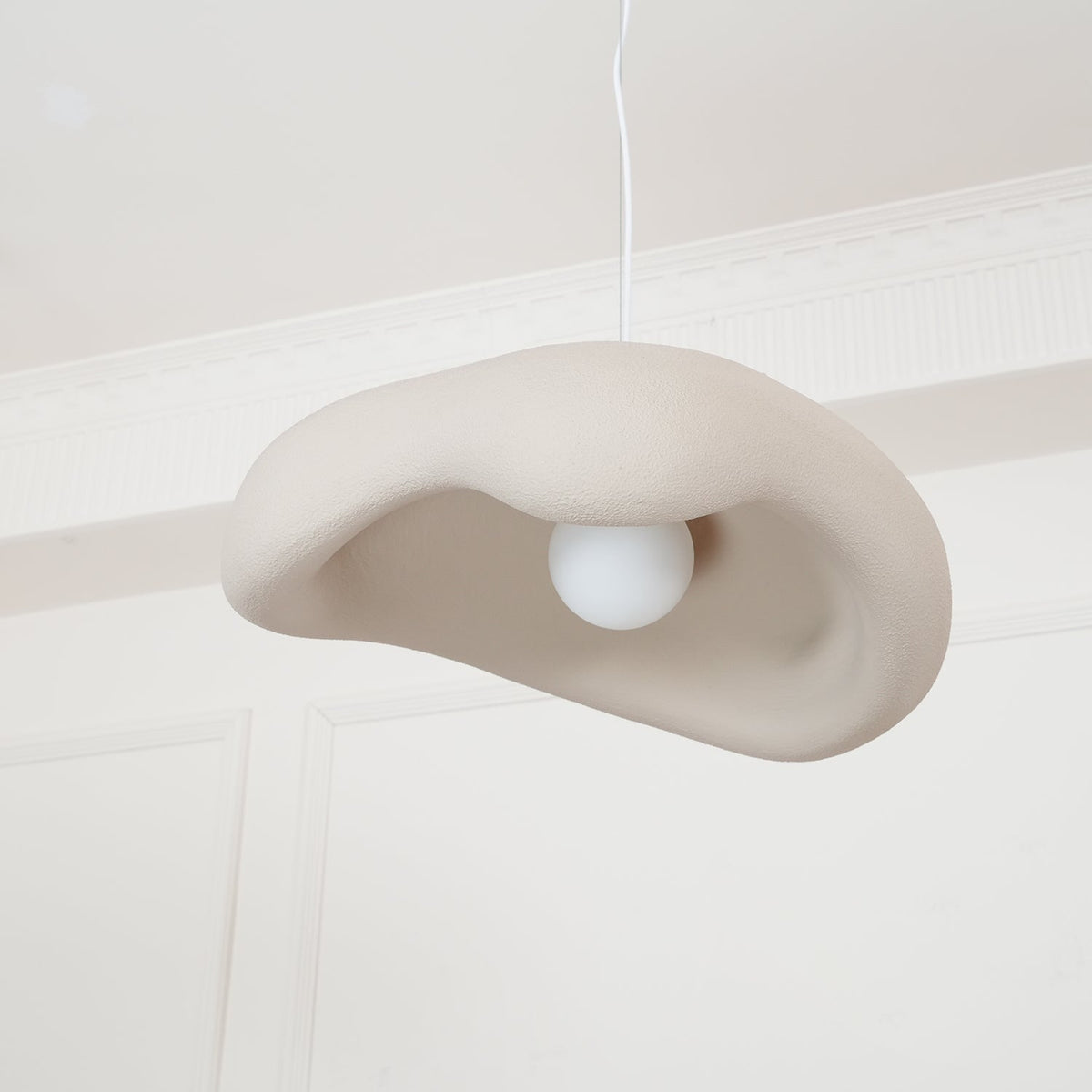 Beige Clérachic pendant seen from low down, with a round white bulb inside the folded shade, under a panelled white ceiling.