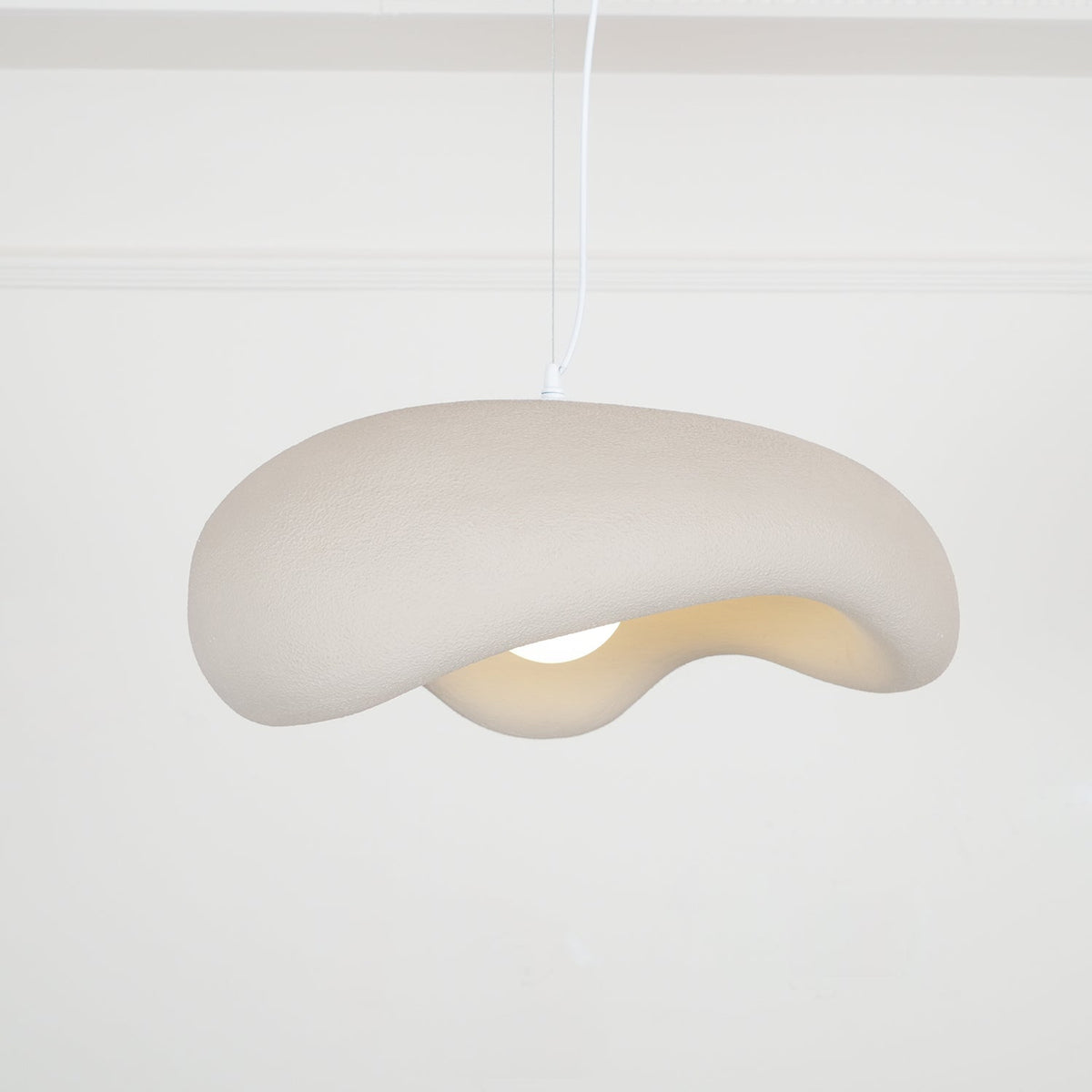 Beige Clérachic pendant seen from below at an angle, its rough shade curling over a bulb, on a white cord against panelled walls.