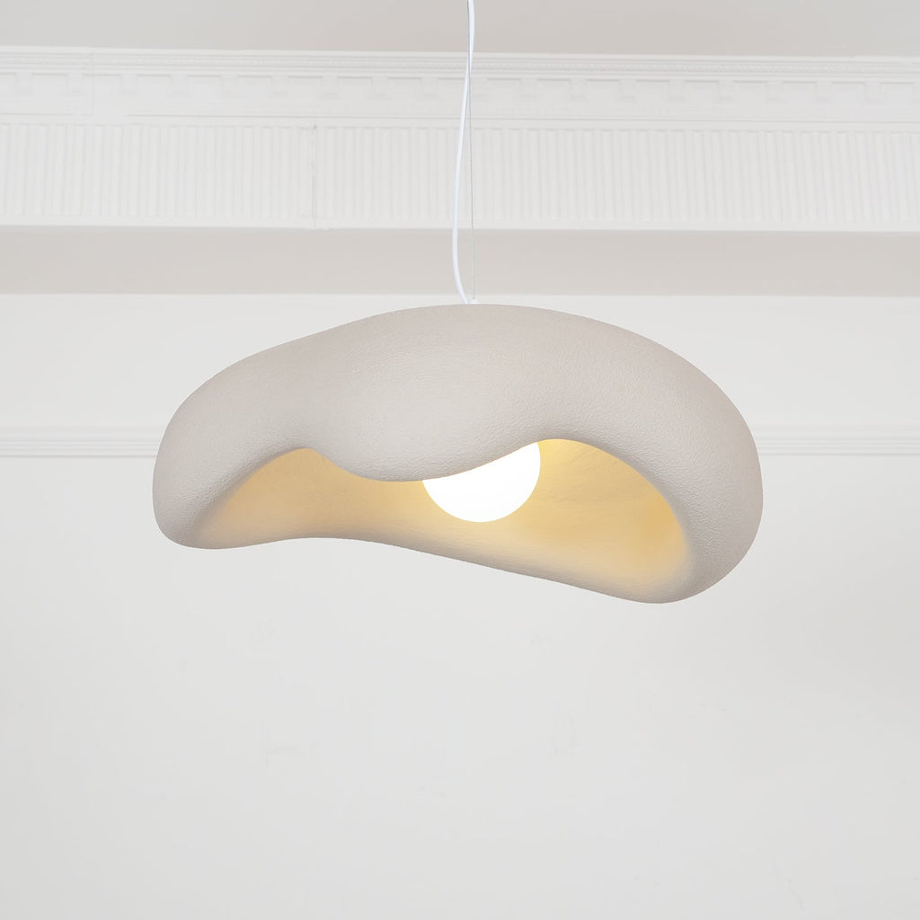 Beige Clérachic pendant hanging in front of a white panelled wall, the wavy shade showing a warm cream interior and a round bulb.