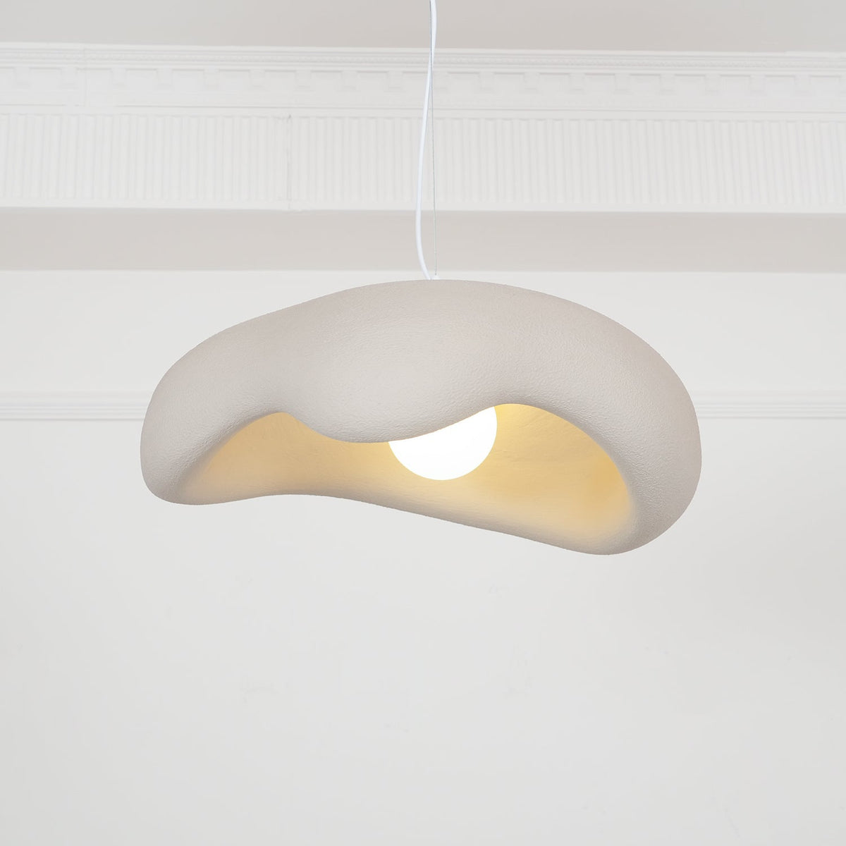 Beige Clérachic pendant hanging in front of a white panelled wall, the wavy shade showing a warm cream interior and a round bulb.