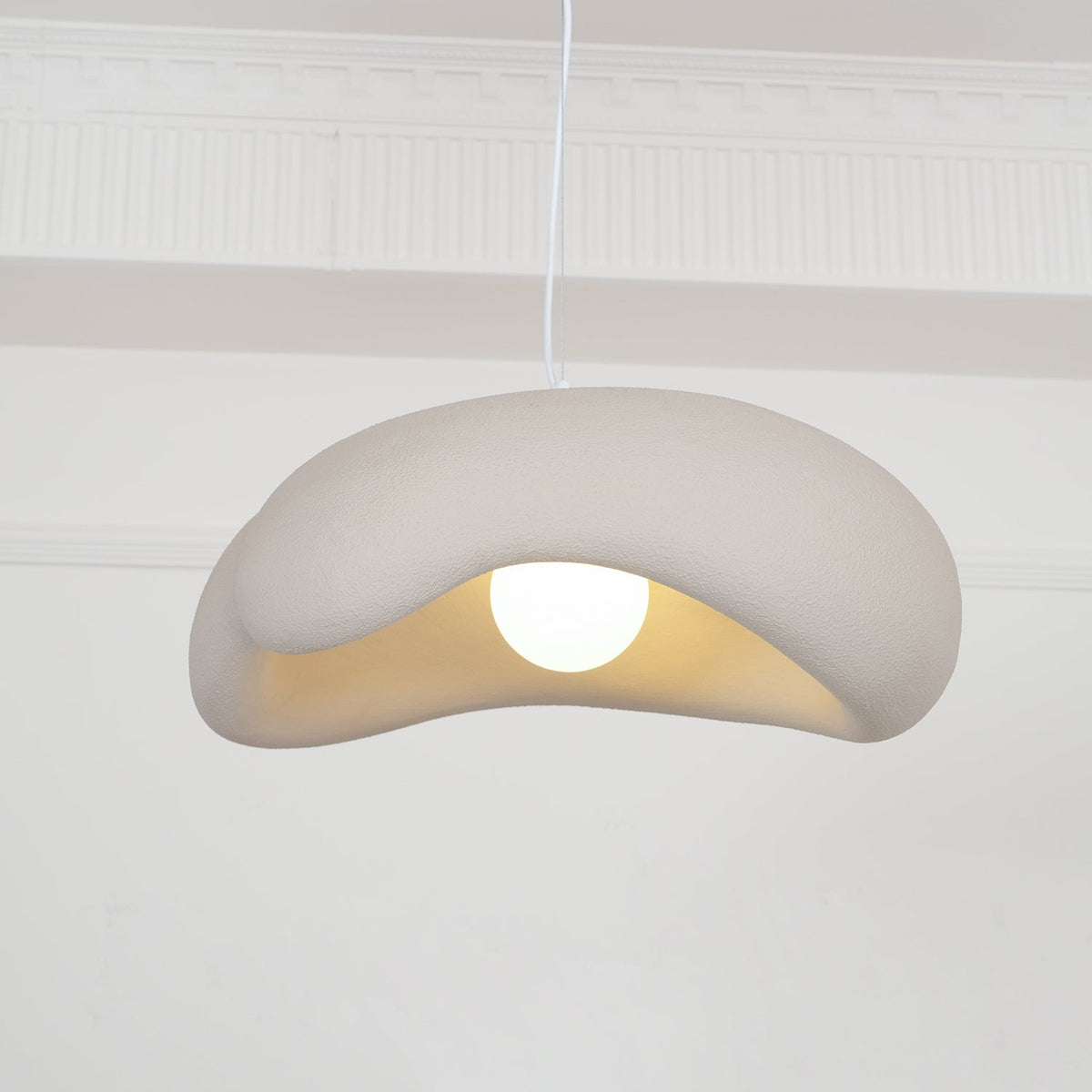 Beige Clérachic pendant with its rolled shade edge and a round bulb, in front of white wall mouldings.