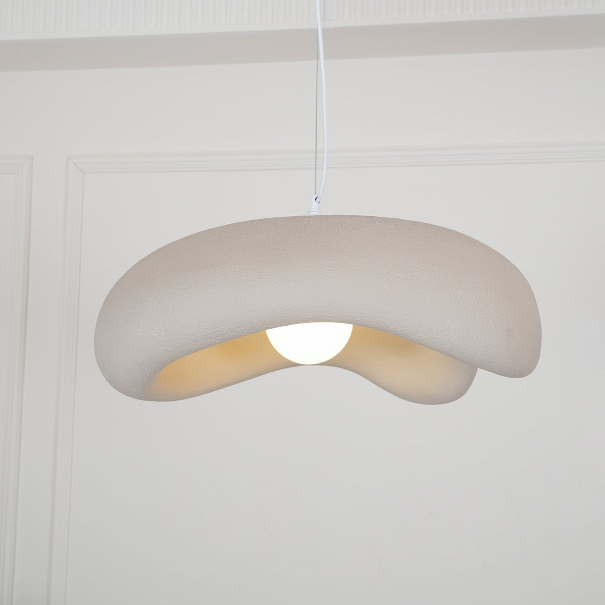 Side view of a beige Clérachic pendant with a thick rolled edge and a round bulb, against white panelled walls.