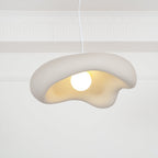 Beige Clérachic pendant with a lit bulb, the textured shade and cream underside close up against a white panelled wall.