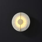 Clérachic round alabaster wall lamp glowing warm from behind its brushed brass centre disc on a charcoal wall.