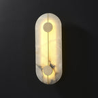 The Clérachic tall alabaster wall lamp lit on a dark wall, its slim brass rod threading two brushed brass discs.
