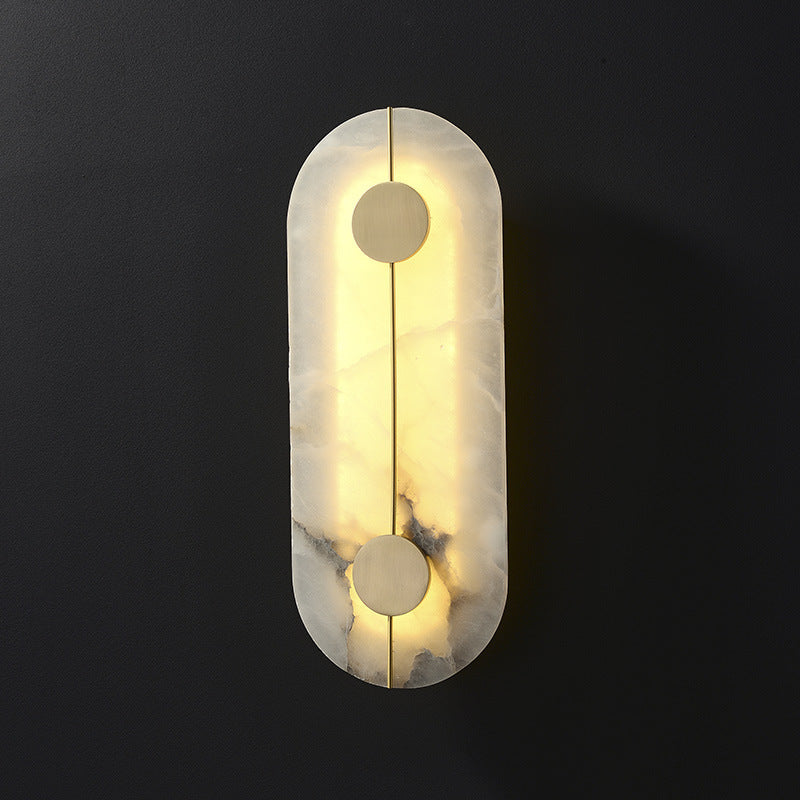 The Clérachic tall alabaster wall lamp lit on a dark wall, its slim brass rod threading two brushed brass discs.