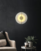Clérachic round alabaster sconce on a black wall above an armchair, the brass hub catching warm light.