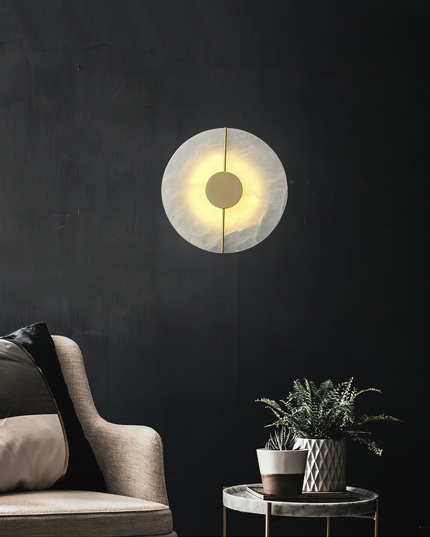 Clérachic round alabaster sconce on a black wall above an armchair, the brass hub catching warm light.