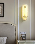 The Clérachic tall alabaster sconce lit on panelled wall beside a bed, with framed art and a clock nearby.
