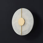 Clérachic round wall lamp unlit against dark grey, the brushed brass disc and slim rod crossing pale alabaster.