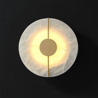 Clérachic round wall lamp lit on black, warm light escaping behind the brass disc and washing the stone.