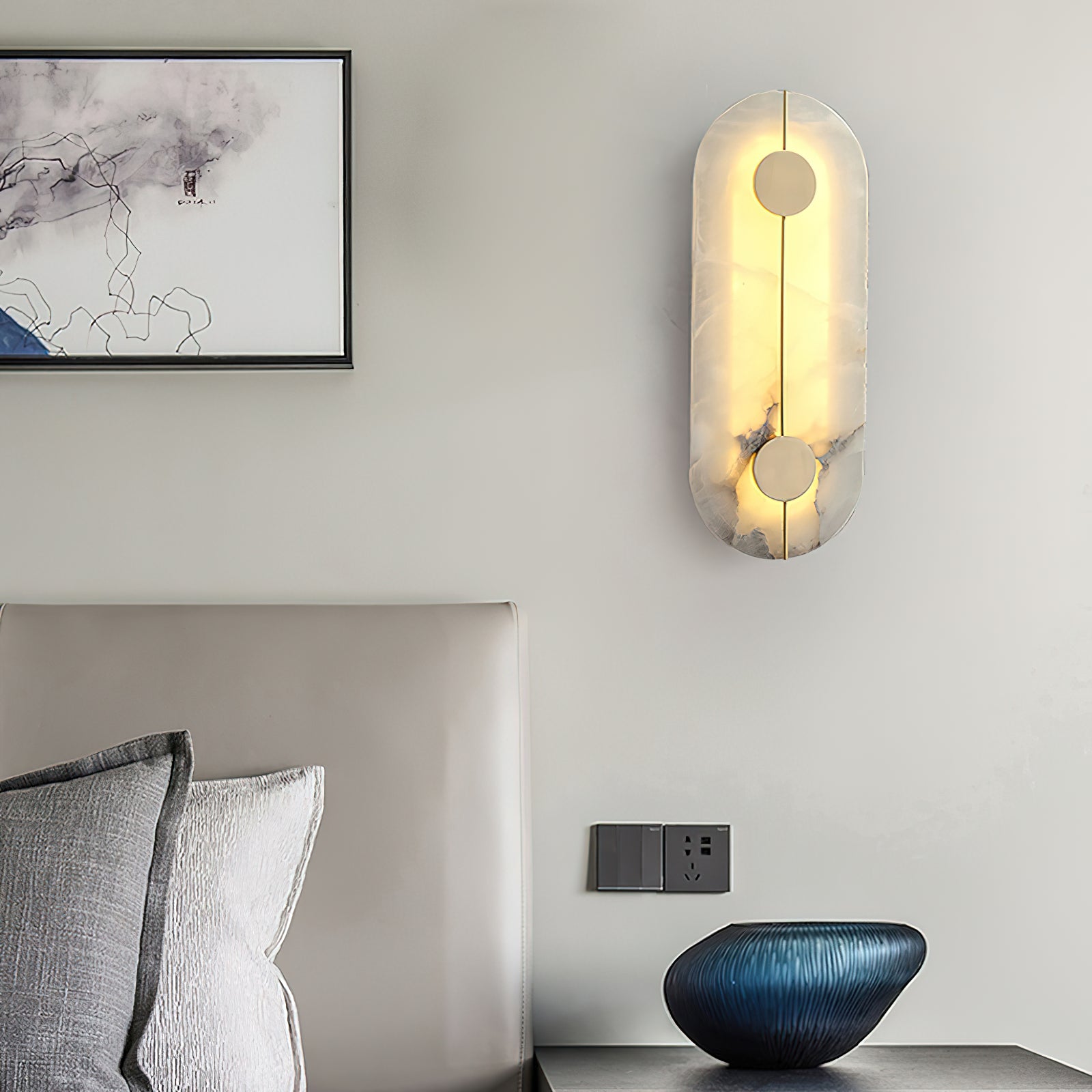 The Clérachic tall alabaster sconce glowing above a bed ledge, a blue vessel and framed print beside it.