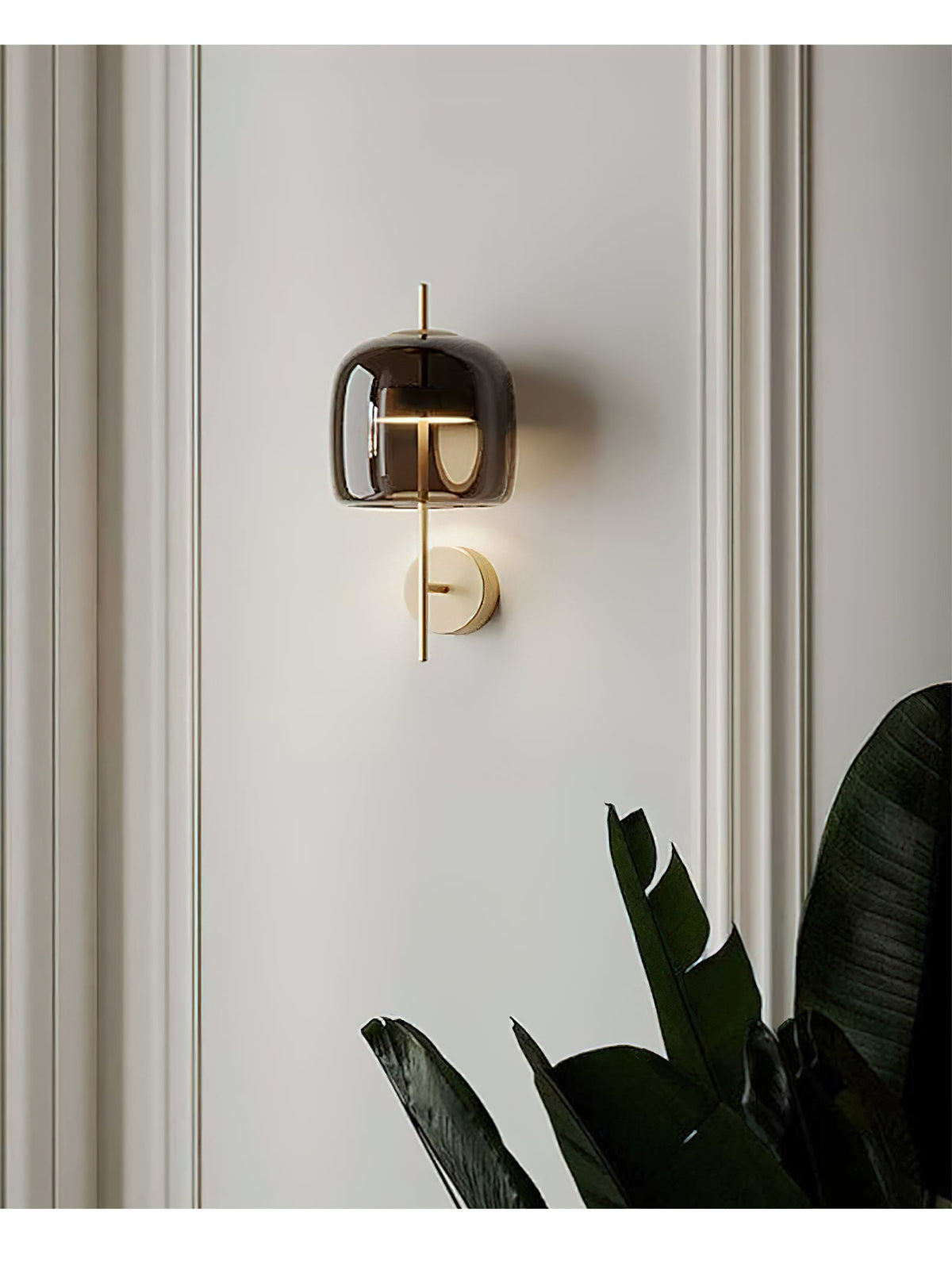 This Clérachic wall light, with a smoke grey shade and gold stem, on a pale panelled wall beside the leaves of a dark green plant.