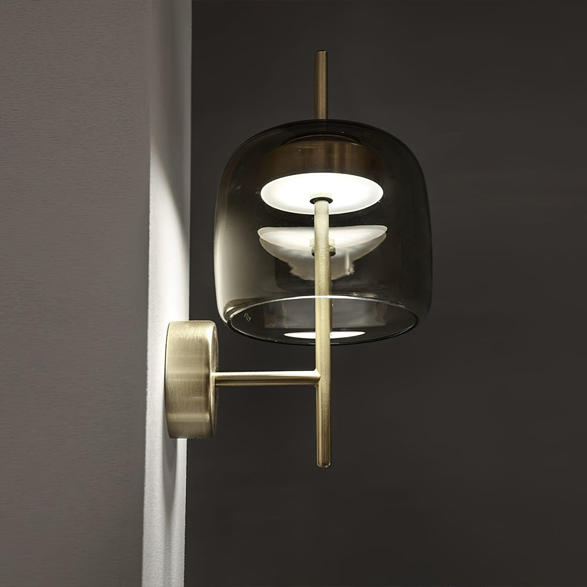 Side view of a Clérachic wall light with a smoke grey glass shade lit from inside, and a brushed gold arm, against a dark wall.