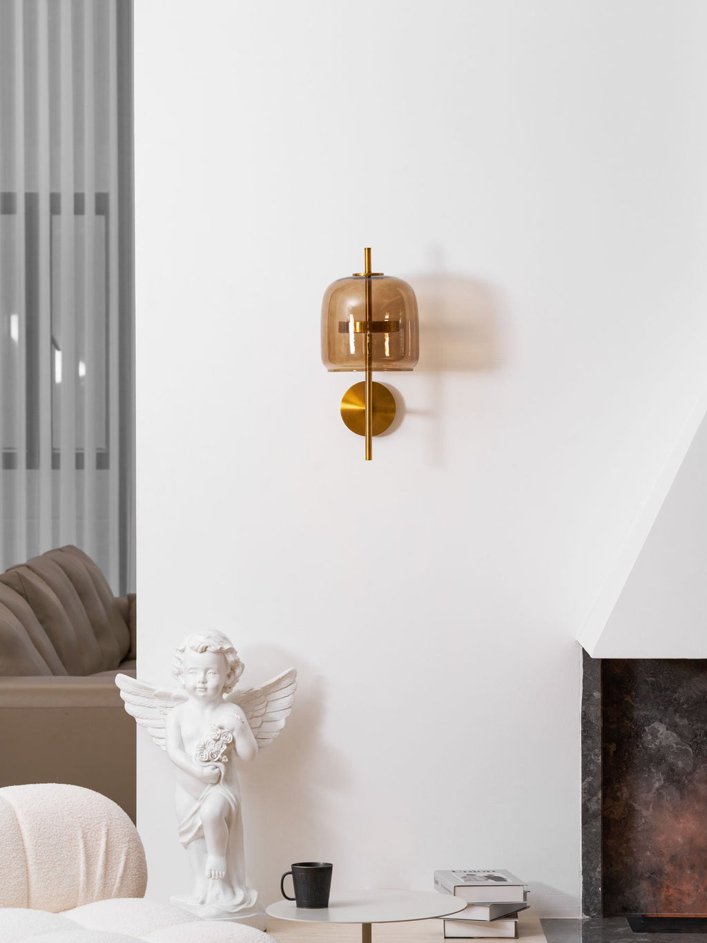 A small Clérachic wall light with a coffee glass shade on a white wall above a cherub statue, a mug and a cream armchair.