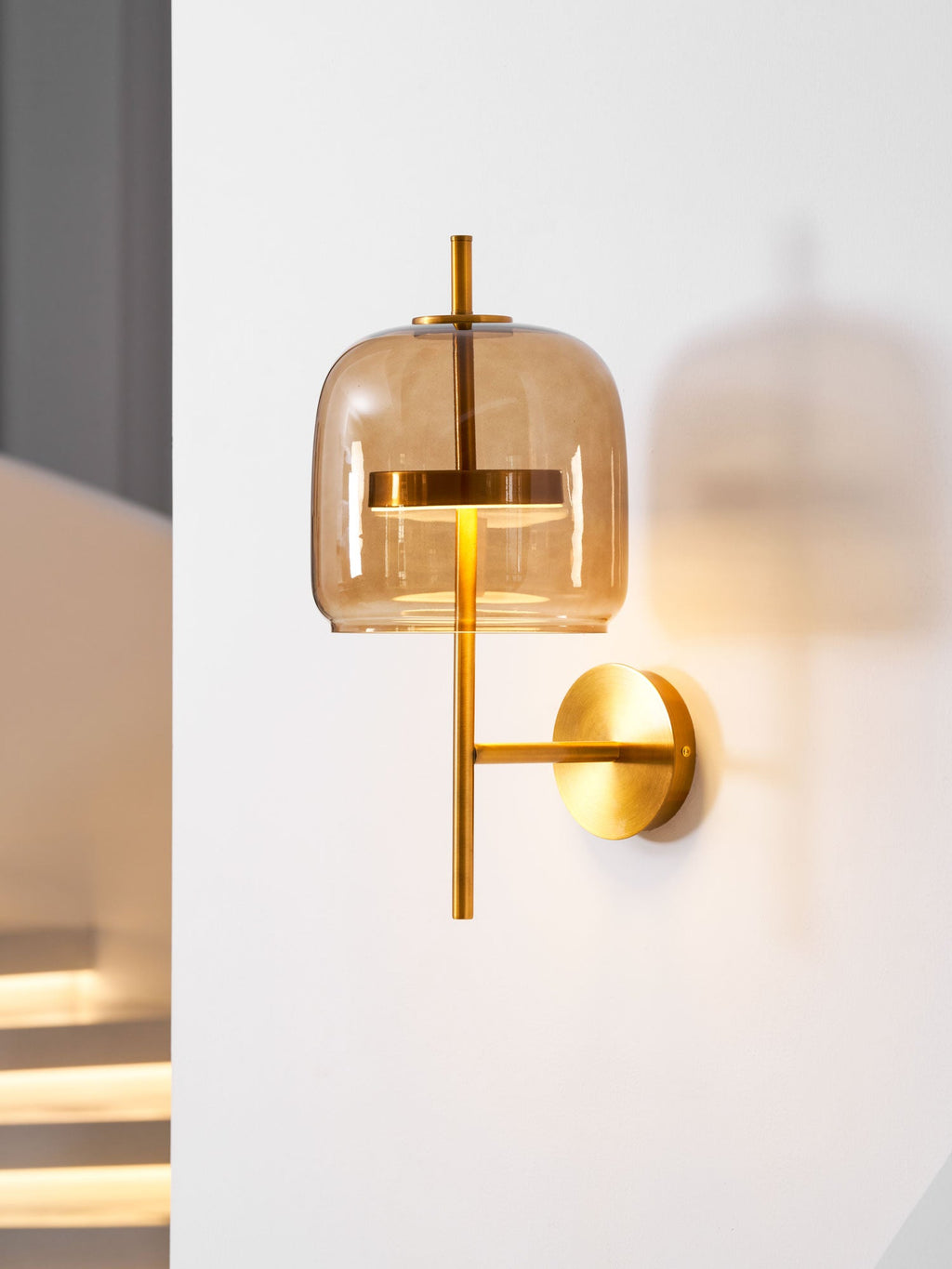 The coffee glass shade of this Clérachic wall light glows softly on a white wall, with lit stair steps blurred behind it.
