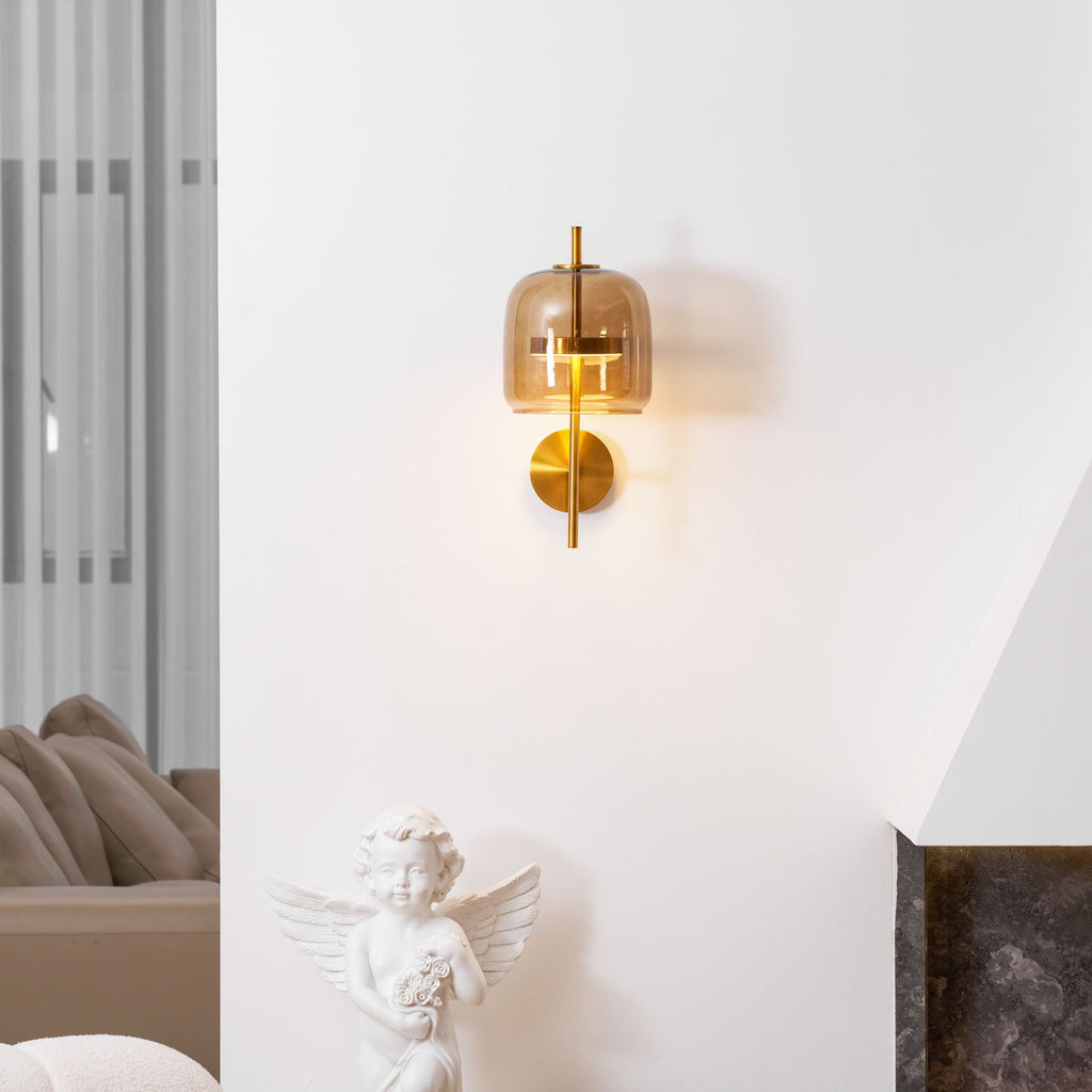 Lit Clérachic wall light with a coffee glass shade and gold stem on a white wall, above a cherub statue and a dark stone fireplace.
