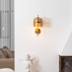 Lit Clérachic wall light with a coffee glass shade and gold stem on a white wall, above a cherub statue and a dark stone fireplace.