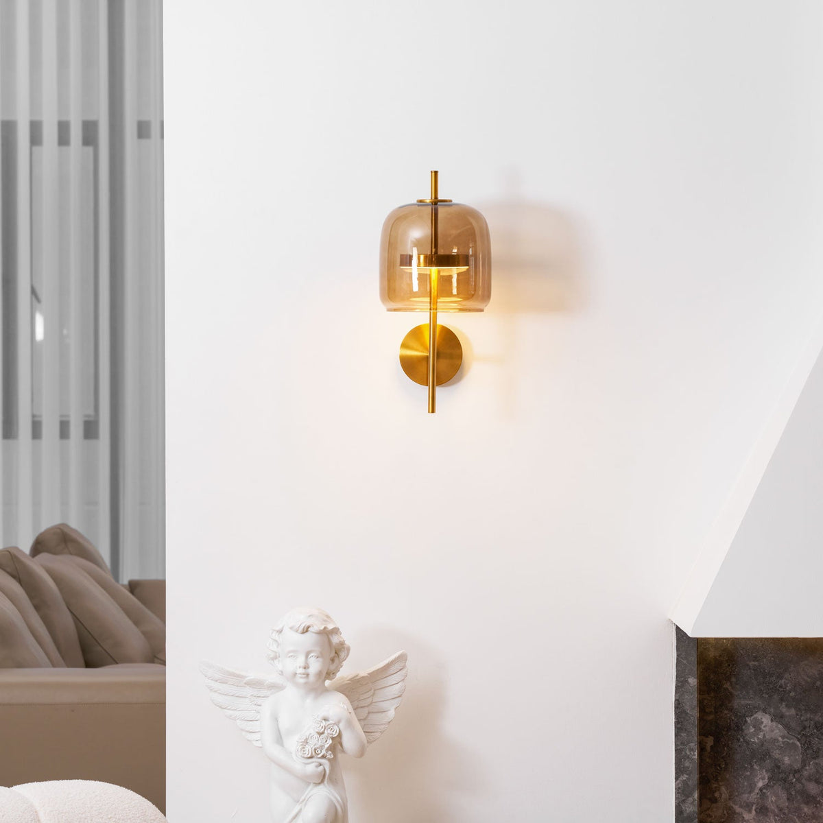Lit Clérachic wall light with a coffee glass shade and gold stem on a white wall, above a cherub statue and a dark stone fireplace.