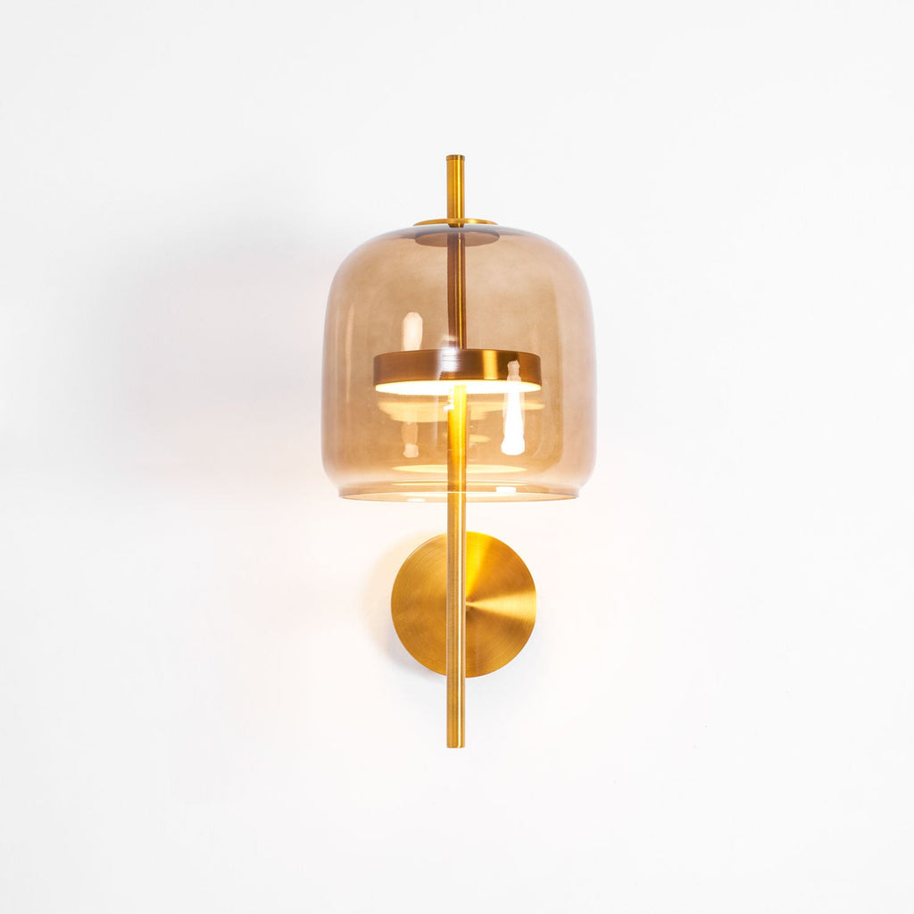 Lit Clérachic wall light with a coffee glass shade, gold stem and round gold backplate, glowing warm on a white wall.