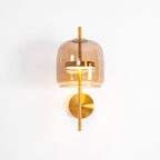 Lit Clérachic wall light with a coffee glass shade, gold stem and round gold backplate, glowing warm on a white wall.