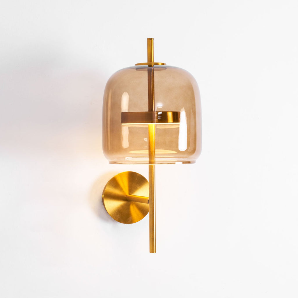 Side-angled view of this Clérachic wall light, with the lit coffee glass shade, gold stem and round backplate on a white wall.