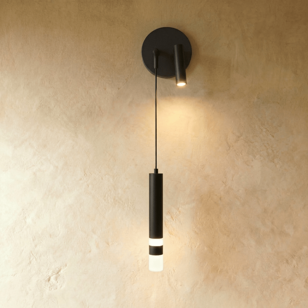 Clérachic matte black wall light with a round backplate, a small spotlight head and a hanging cylinder with a lit frosted band.