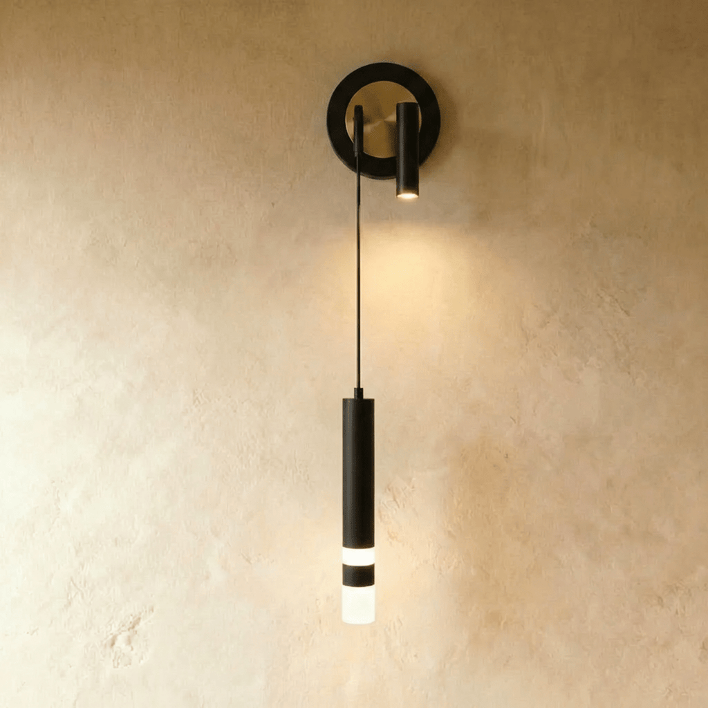 Clérachic wall light with a black ring backplate framing a brass-toned disc, a spotlight head and a black hanging cylinder.