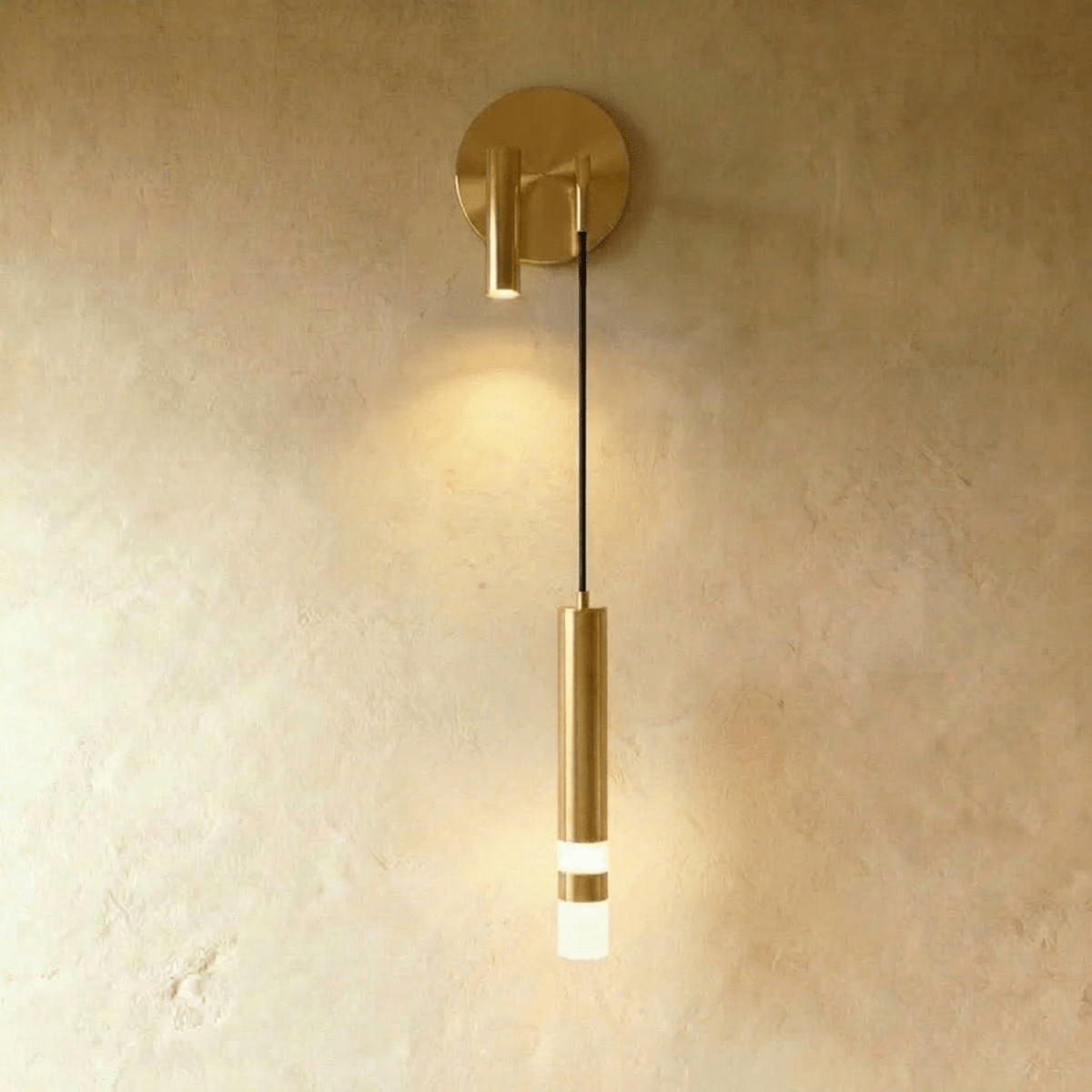 Clérachic brass-toned wall light with a round backplate, a spotlight on the opposite side and a lit hanging cylinder.