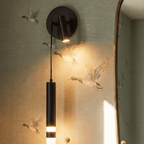 Close view of a Clérachic matte black wall light, its spotlight lighting flying-bird wallpaper above a glowing frosted cylinder.