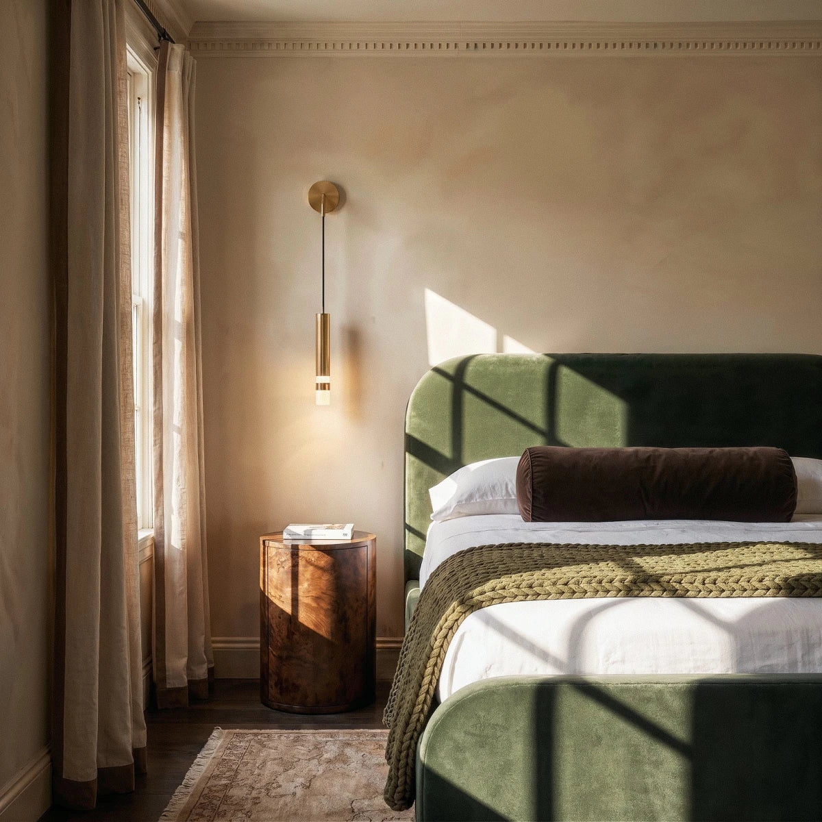 Clérachic brass-toned wall light with a slim cord and glowing cylinder beside a green velvet bed in a warm, sunlit bedroom.