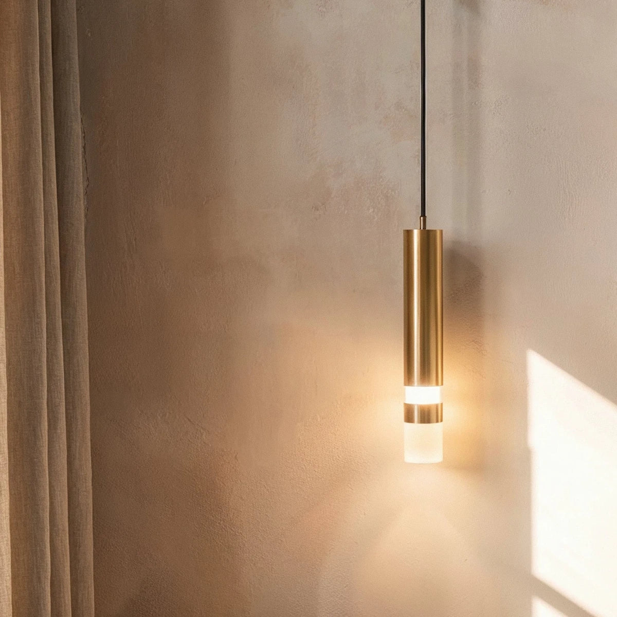 Clérachic brass-toned cylinder wall light with a frosted glass tip, glowing beside a linen curtain on a limewashed wall.