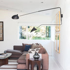 Black Clérachic wall light with long arms reaching across a white living room, above a plum sofa near a large window.