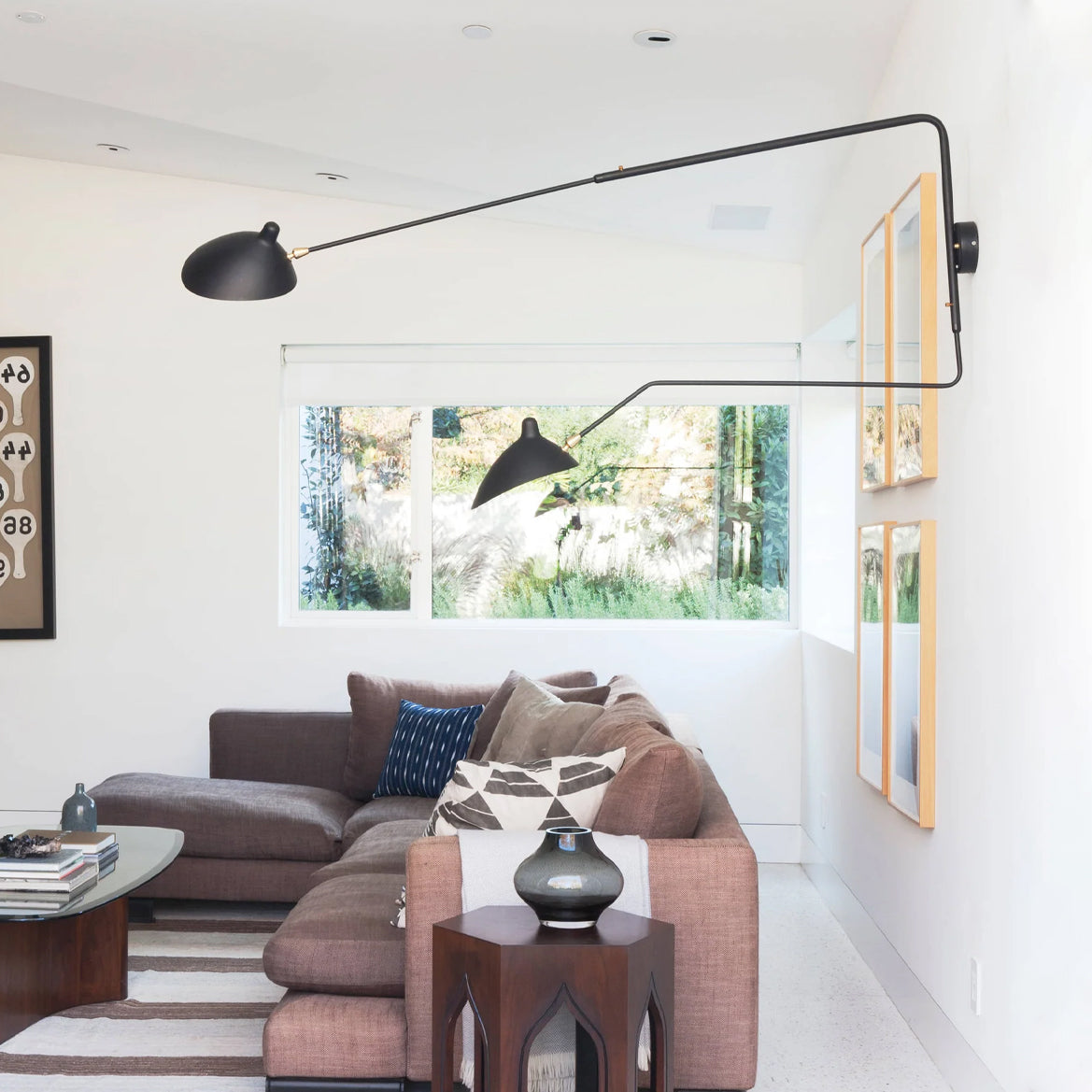 Black Clérachic wall light with long arms reaching across a white living room, above a plum sofa near a large window.