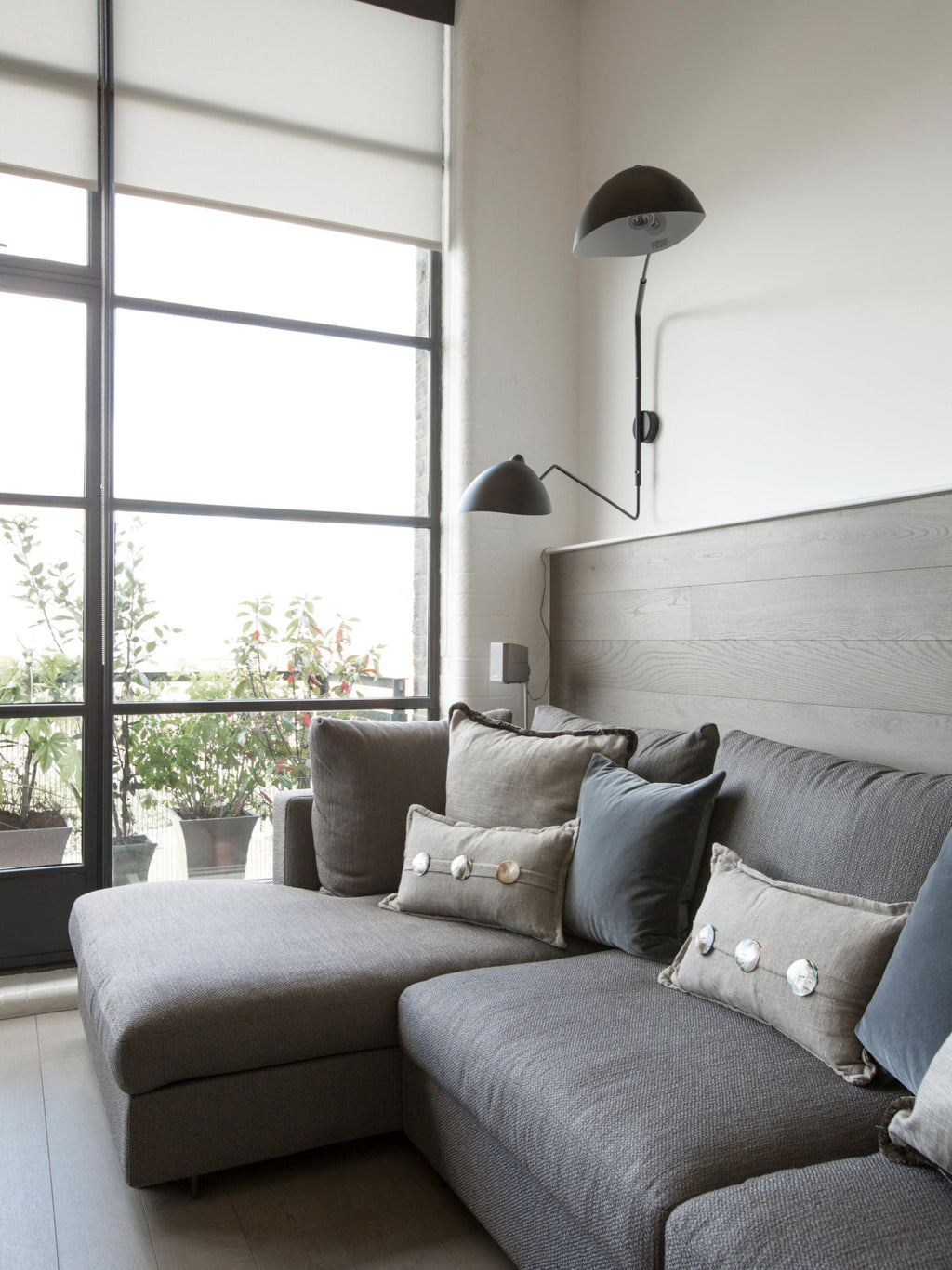 Black Clérachic wall light with a long curved arm beside a tall window, above a grey corner sofa with scatter cushions.