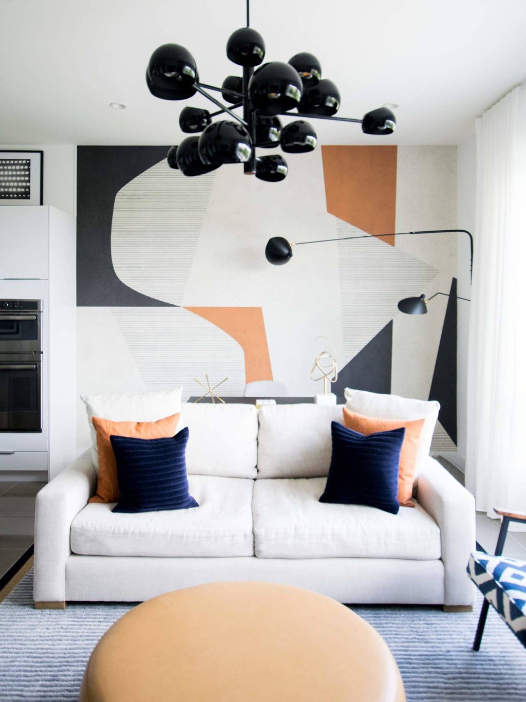 Clérachic black wall lights on an abstract mural wall, above a white sofa with orange and navy cushions and a cluster of black globe lights.