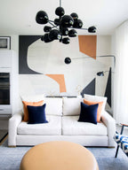 Clérachic black wall lights on an abstract mural wall, above a white sofa with orange and navy cushions and a cluster of black globe lights.