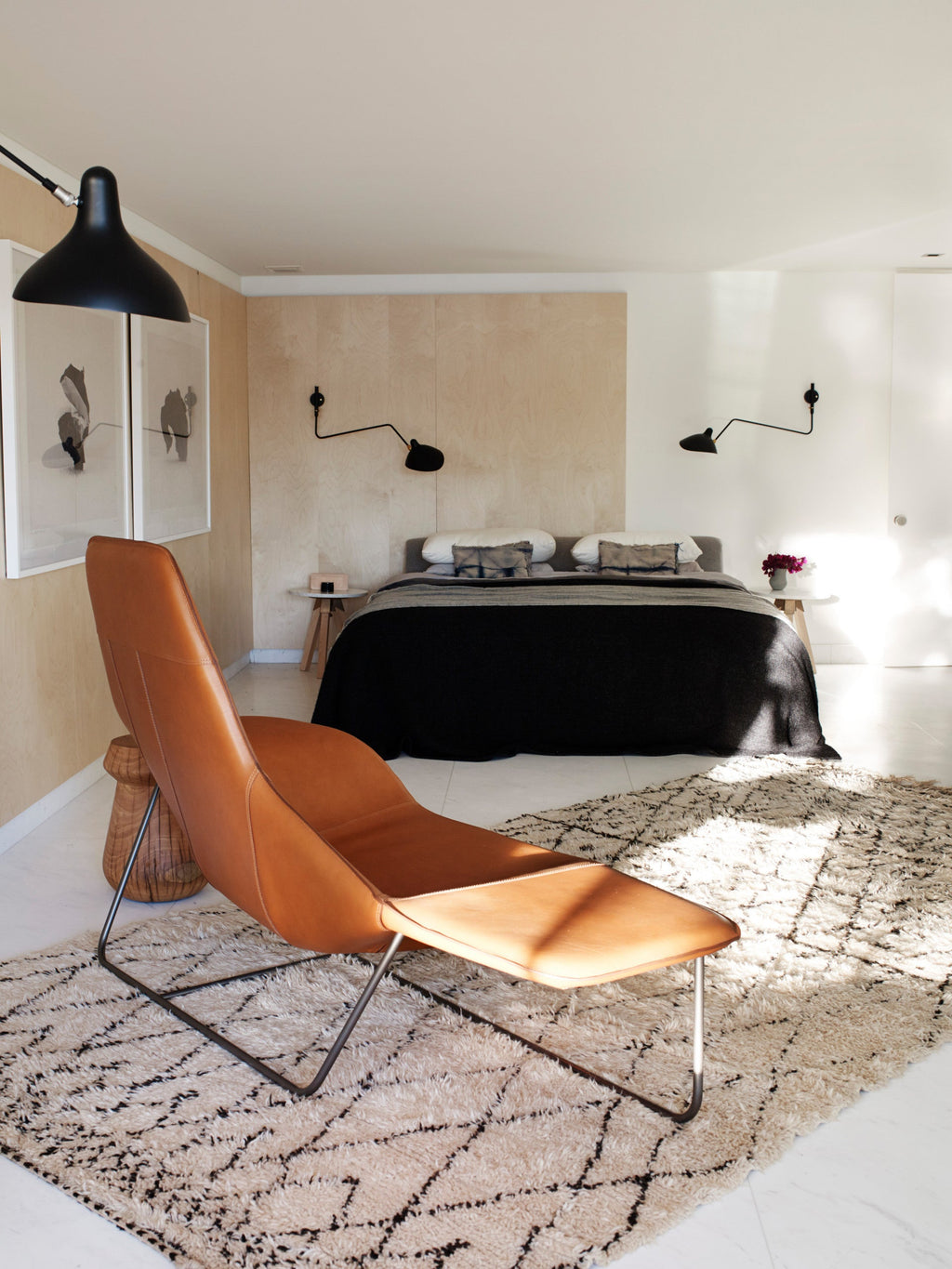 A black Clérachic wall light on a pale wood wall above a bed, with a tan leather lounge chair on a patterned rug.