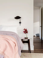 A small black Clérachic wall light with a short arm above a bed with a pink throw and a round side table with red flowers.