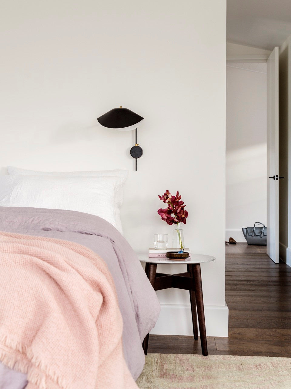 A small black Clérachic wall light with a short arm above a bed with a pink throw and a round side table with red flowers.