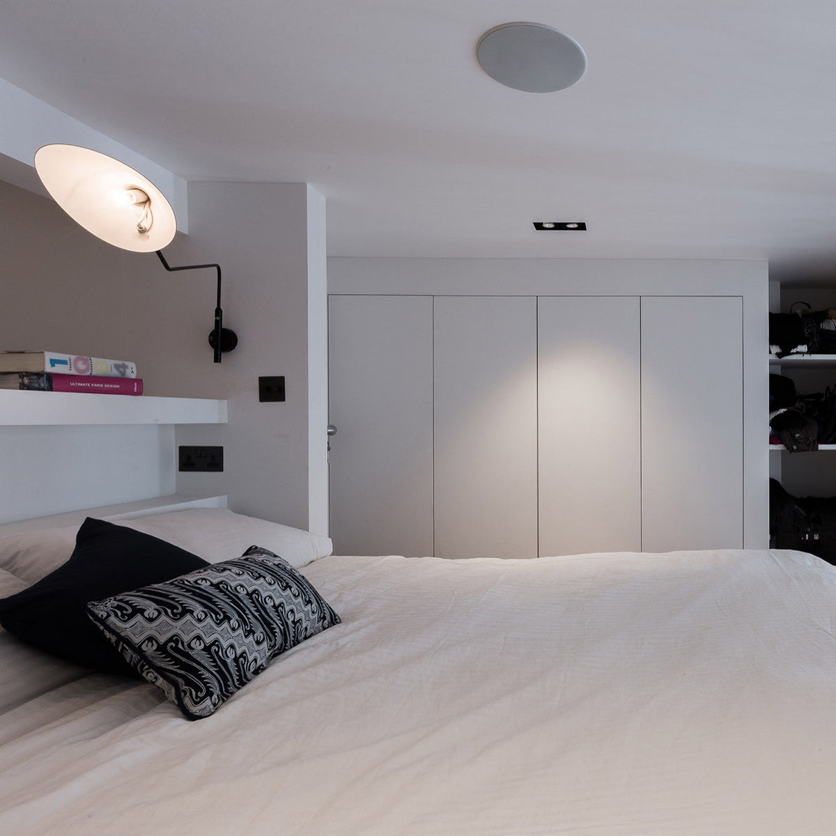 Clérachic wall light with a cone head angled up, lit, above a white bed with a patterned cushion, in a white bedroom with built-in wardrobes.