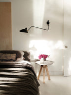 A single Clérachic black wall light with a long arm above a bed, on a white wall beside a round side table with pink flowers.