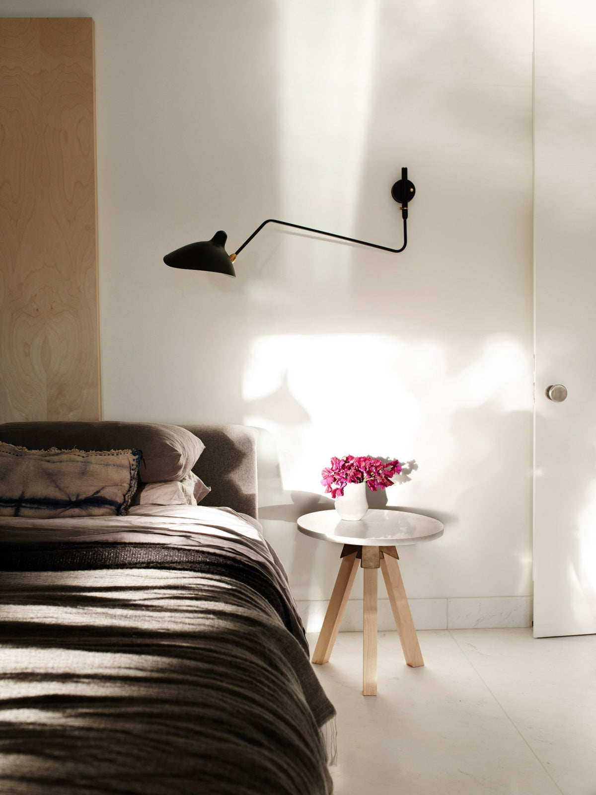A single Clérachic black wall light with a long arm above a bed, on a white wall beside a round side table with pink flowers.