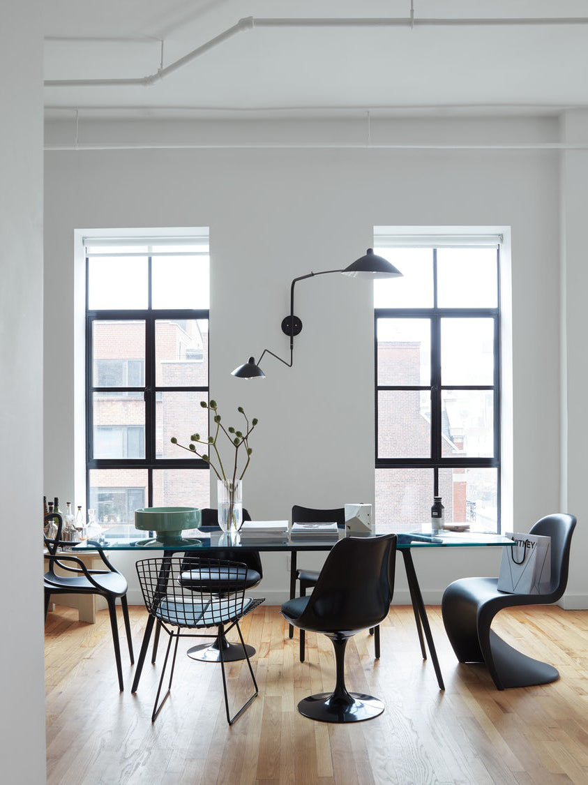 Clérachic black wall light with two long arms beside tall windows, above a dining table with black chairs.