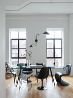 Clérachic black wall light with two long arms beside tall windows, above a dining table with black chairs.