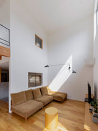 Clérachic wall light with long black arms in a tall living room with pale walls, above a tan sofa and a round yellow stool.