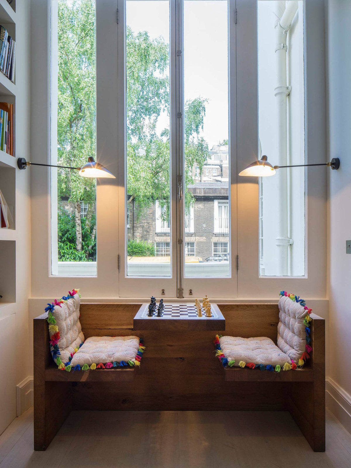 Pair of Clérachic black wall lights flanking a tall window, above a wooden bench with a chessboard between the seats.