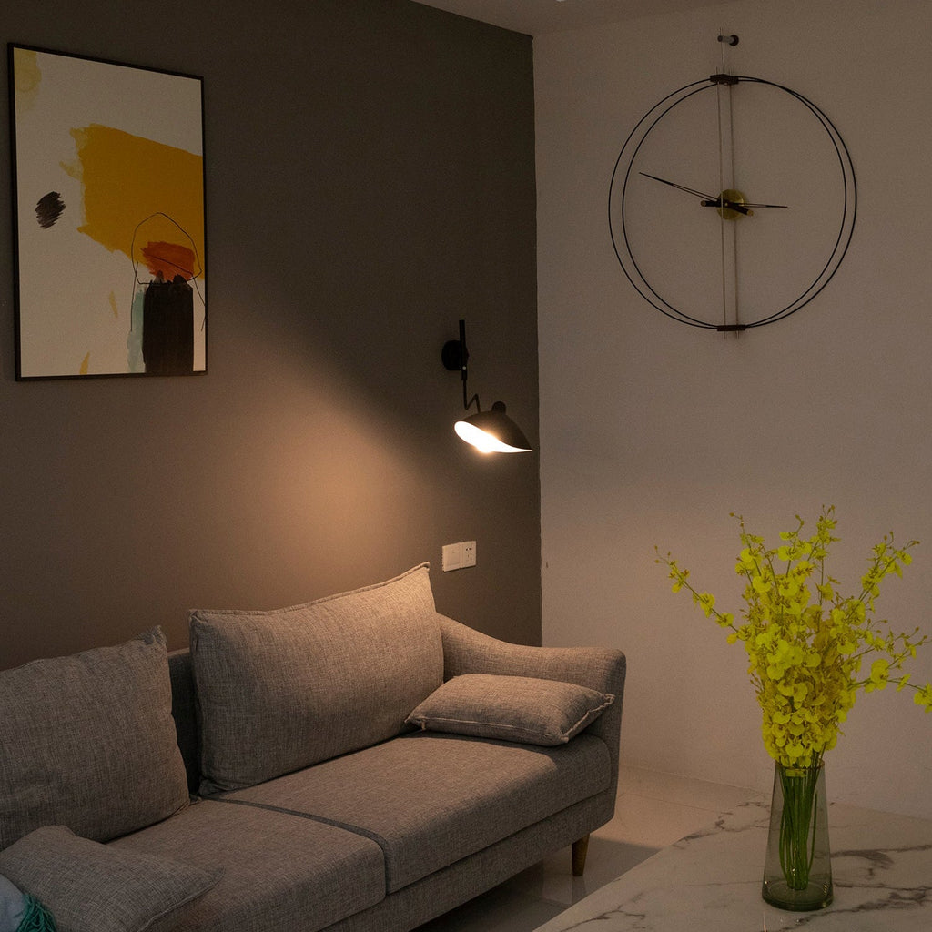 Lit Clérachic black wall light with a small shade above a grey sofa, on a dark wall beside a large round clock.
