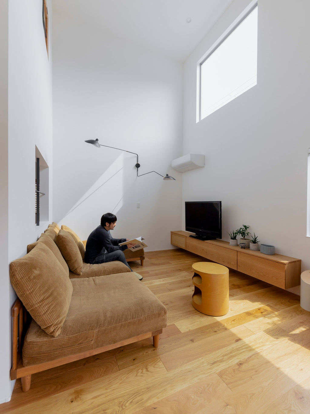Clérachic wall light with two long arms above a man seated on a low tan sofa, in a tall white room with a wooden TV unit.