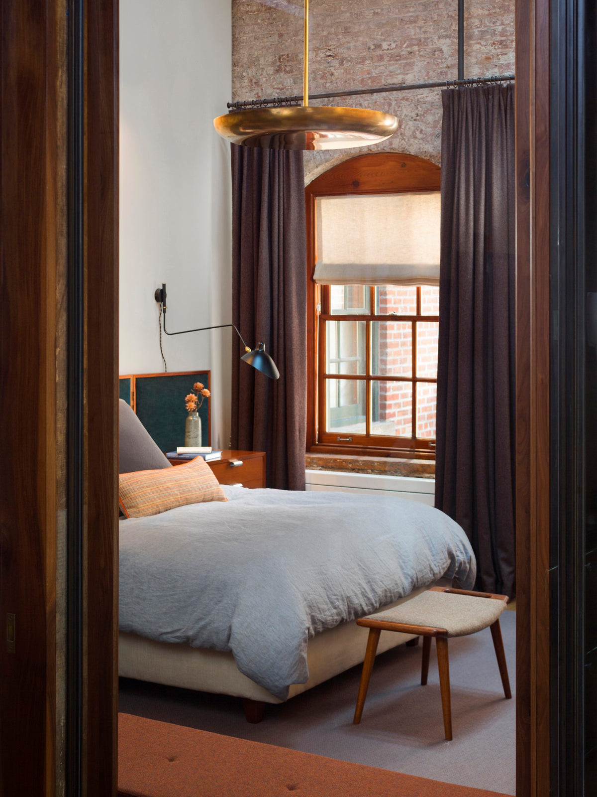 A small Clérachic wall light on a white wall by an arched window with dark curtains, in a bedroom with a pale blue bed.